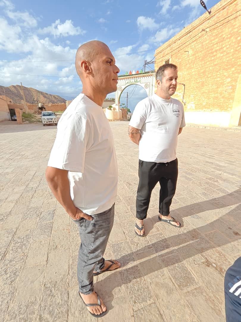 Due viaggiatori su una piazza lastricata di paese davanti a un arco cerimoniale dipinto