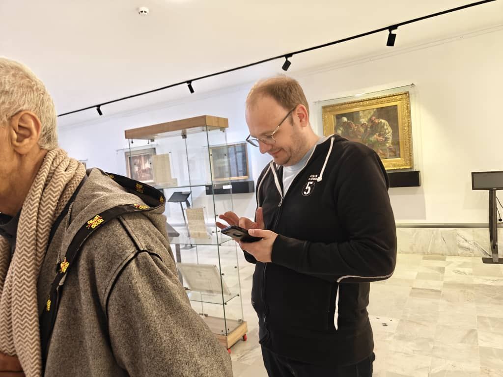 Visitatori leggono le didascalie in una sala di museo con vetrine e dipinti incorniciati