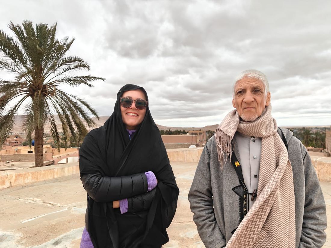 Un viaggiatore con uno scialle nero e un anziano del posto su una terrazza, con palme e paese sullo sfondo