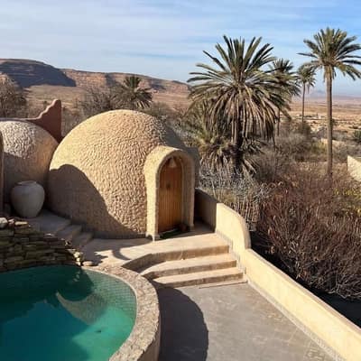 Domed earthen guest lodgings and a small pool at Bou Saâda, palms and desert hills beyond