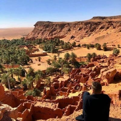 A traveller looking over a ruined ksar and palm grove below the cliffs of the Gourara