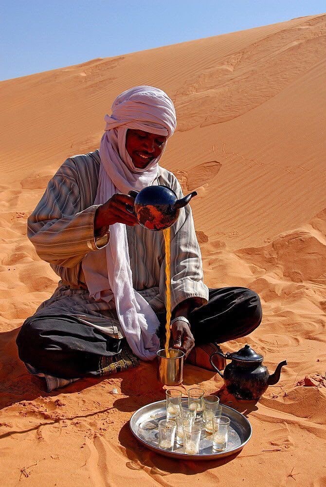 A Tuareg man in a white turban pouring tea from a height on the dunes