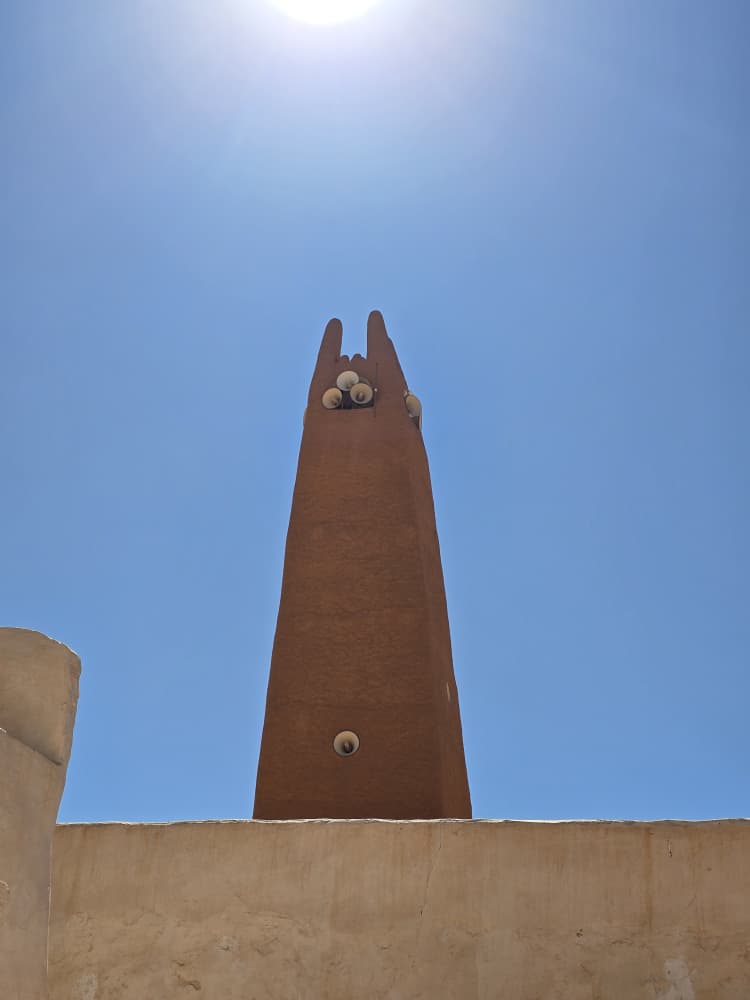 The plain ochre minaret of a M'Zab mosque rising into a deep blue sky, its four finials against the sun